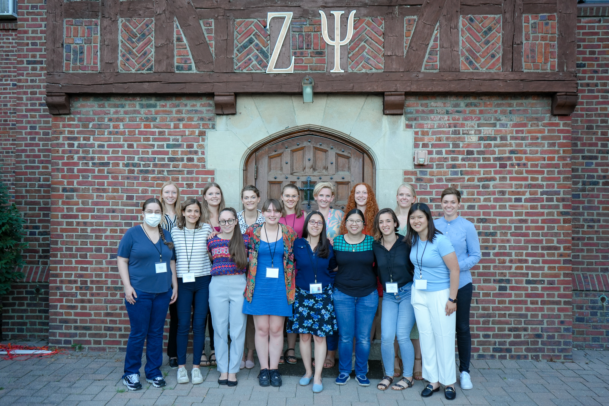 A group of students stand in two rows and smile for the camera outside a brick fraternity house