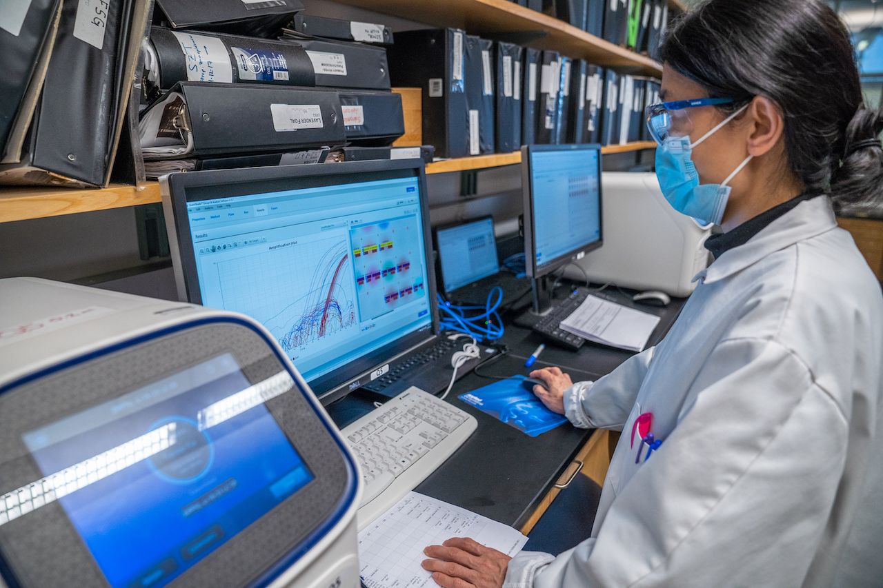 A masked woman in a lab coat and gloves examines a monitor in a lab