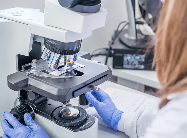 A woman in lab coat and gloves looking into a microscope