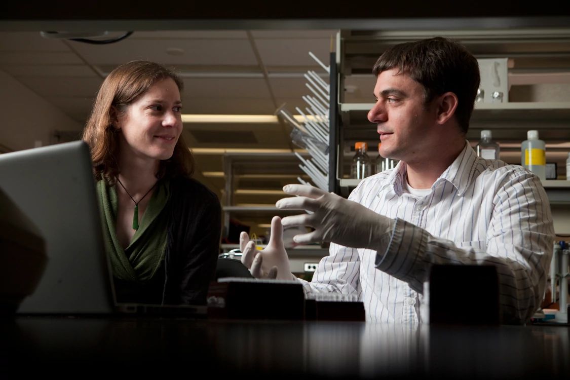Adam Boyko talks to a female postdoc while sitting in the lab behind a laptop