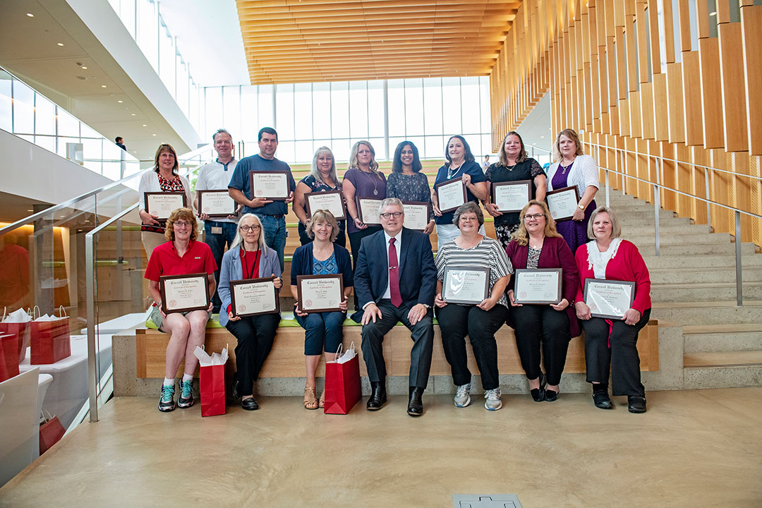 Staff service award recipients sit with Dean Warnick and their plaques on the steps in the college atrium