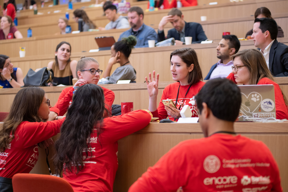 A group of students wearing red Animal Health Hackathon shirts are seated in Lecture Hall 4, brainstorming as a team for the event