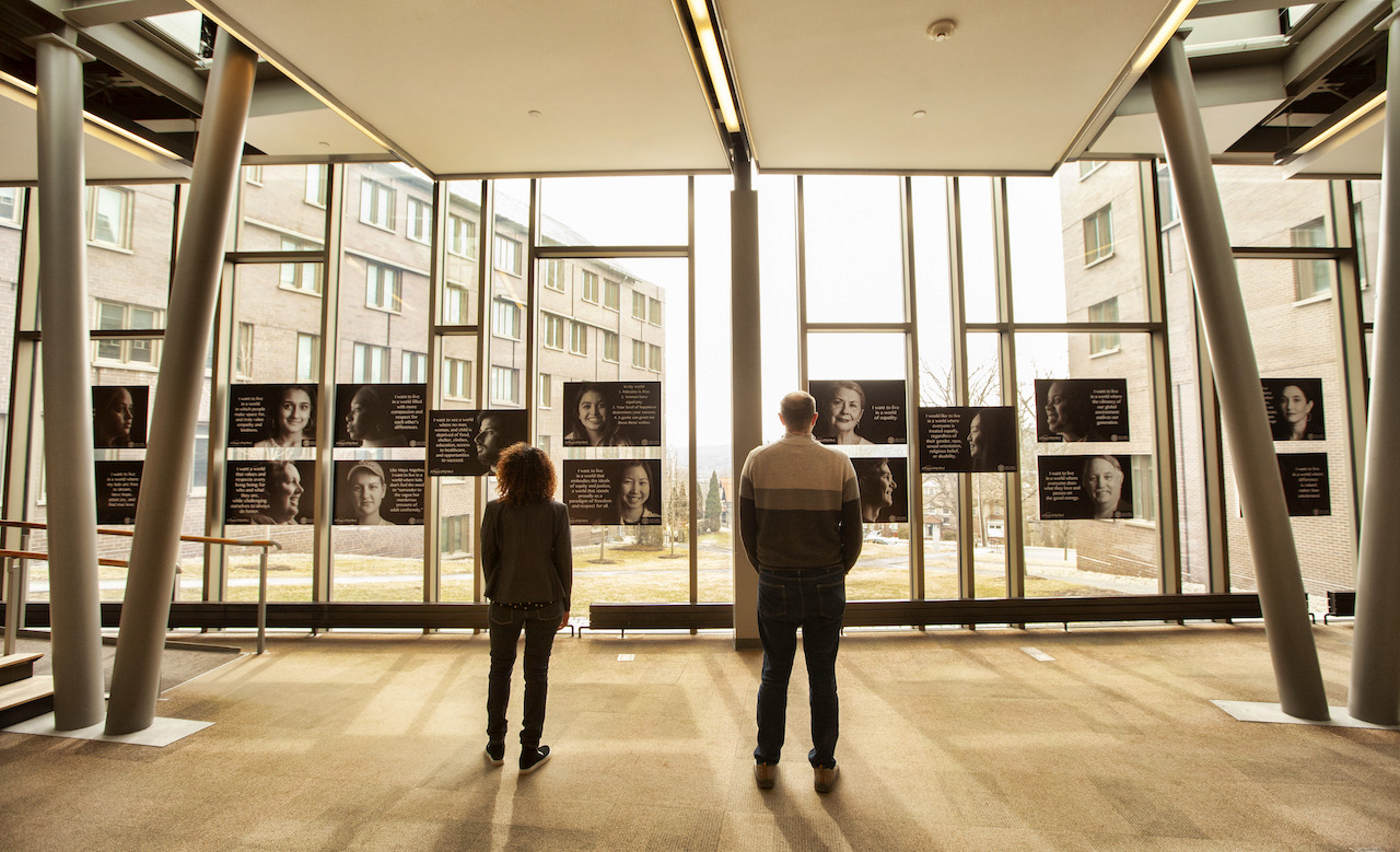 Two people viewed from behind, looking at a photo exhibit displayed on tall, bright windows