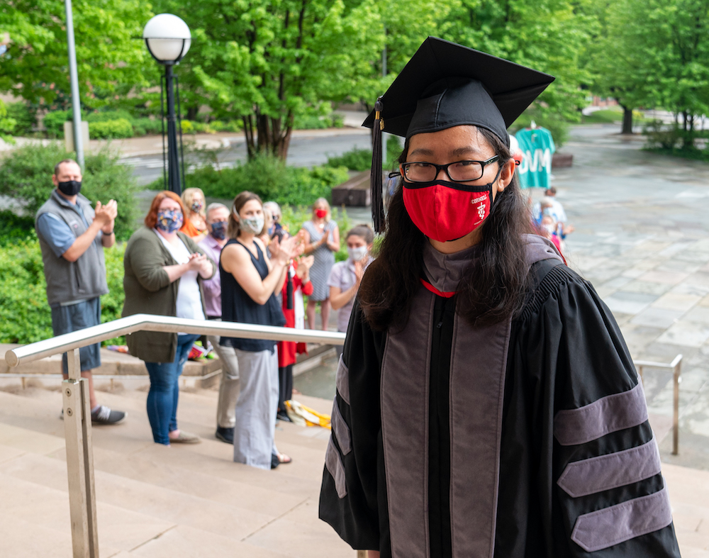 A veterinary student in cap and gown with a Cornell red mask on walks into Bailey Hall, cheered by masked spectators