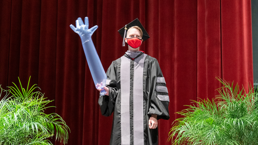 A veterinary student in cap and gown waves an inflated palpation glove on stage at the college's send-off ceremony