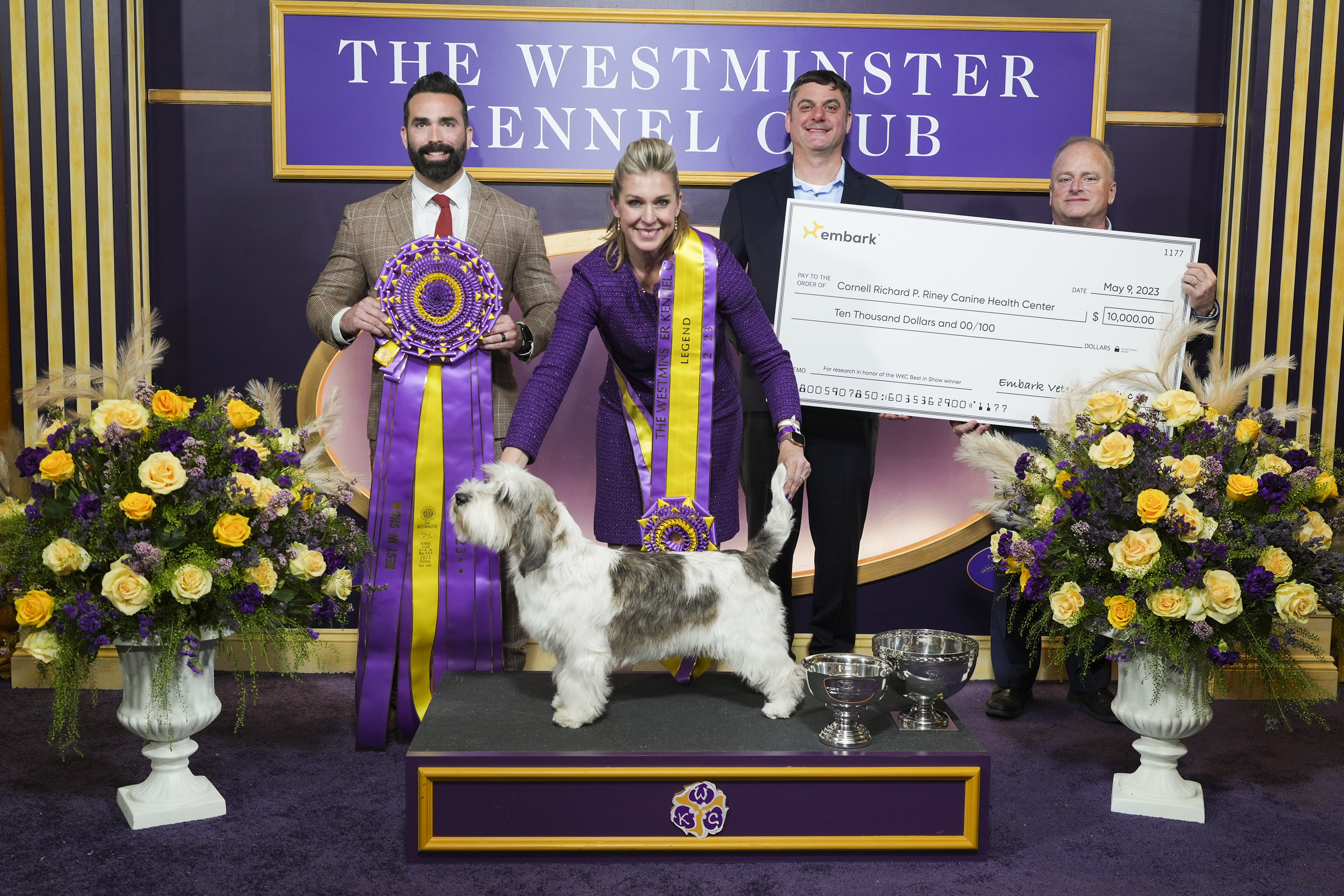 Two men in suits hold a giant check behind a woman and a dog who won giant purple and yellow ribbons