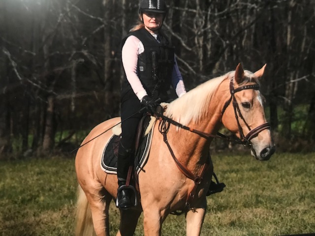 A woman in a vest and helmet rides a palamino horse. 