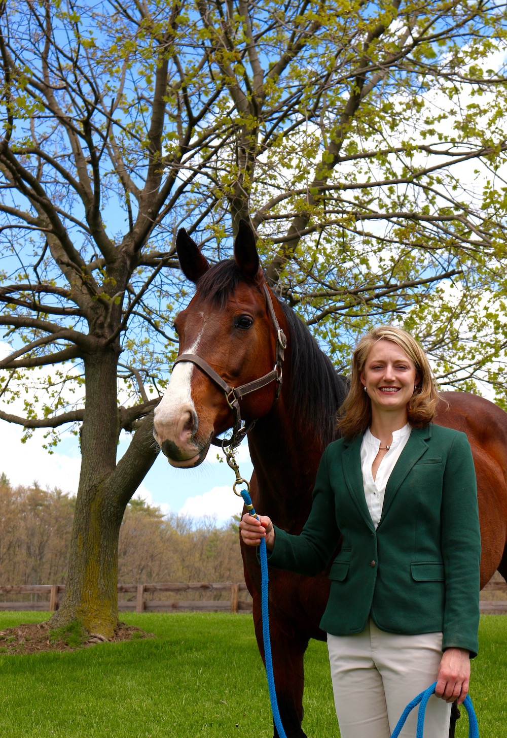 Heidi Reesink, a pale woman with blonde hair, stands next to a brown mare at the Cornell Equine Park; a barn is behind them and it is a bright summer day