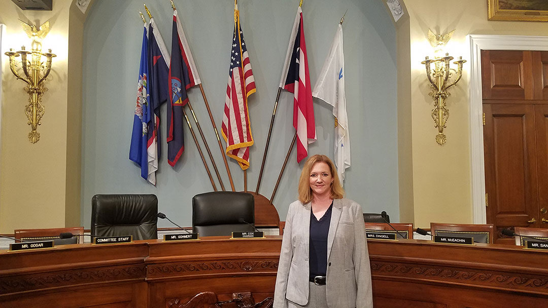 Krysten Schuler standing inside the House of Representatives, with flags and a giant desk behind her