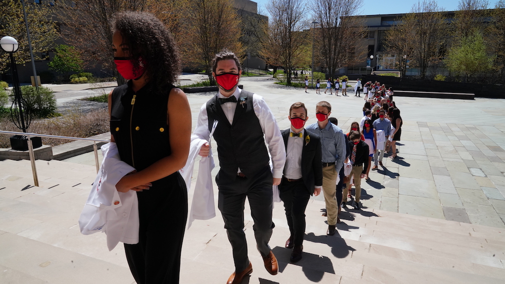 A line of students wearing masks and carrying their white coats into Bailey Hall
