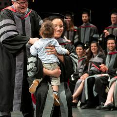A graduate holding a baby receives her hood from Dean Warnick