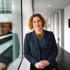 A woman with curly brown hair and a warm smile is standing in a modern office hallway