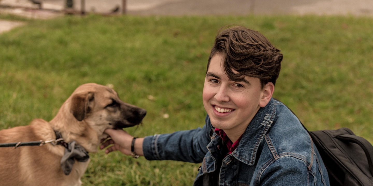A person with dark hair kneels next to and pets a brown dog. 