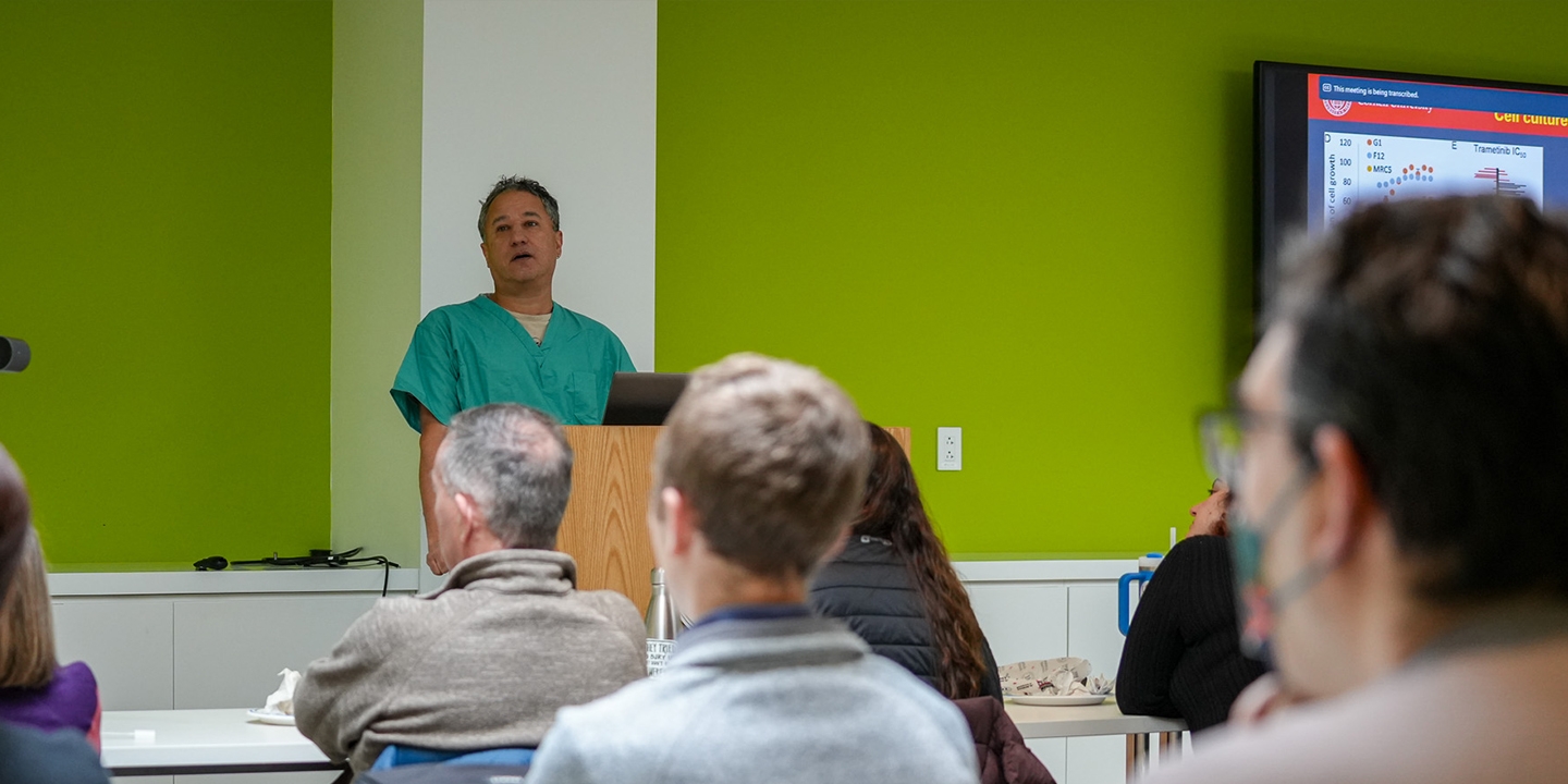 A man in scrubs stands at the front of a room speaking. 