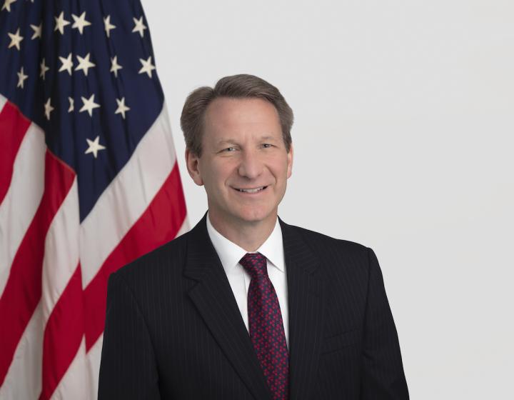 A headshot of Dr. Normal Sharpless, wearing a suit and tie with an American flag backdrop