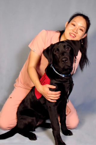 A woman in salmon colored scrubs poses with a black lab on a gray background. 