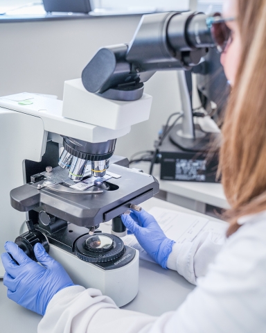 A woman in a lab coat with gloves looks into a microscope