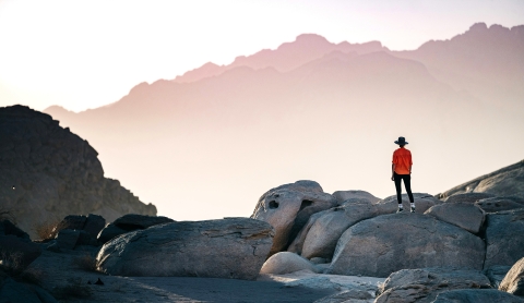 Person in a red jacket standing on rocky terrain, looking out at mist-covered mountains