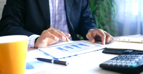 Person in business attire using a tablet with bar charts on a desk with a calculator, notebook, pen, and coffee mug.