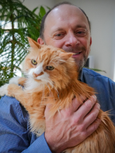 A man with facial hair wearing a blue shirt and holding an orange cat. 
