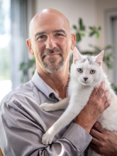 A bald man with a goatee poses with a white cat. 