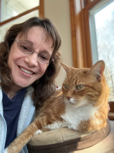 A women with dark hair and glasses poses with a cat while wearing a lab coat. 