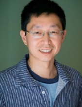 Chun Han in a green background with a blue and white striped collared shirt and wearing metallic thin-framed glasses.