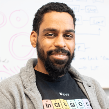 Karl Lewis wearing a chemistry novelty T-shirt and a grey zip-up hoodie in front of a white board with biochemical diagrams.