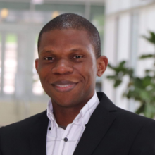 Zeribe Nwosu in a black blazer and white collared shirt with grey squares behind an entrance.