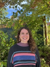 Sydney Womack wearing a knit sweater standing in front a building surrounded by green vegetation.