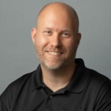 Nicolas Buchon wearing a black polo shirt in front of a gray background.