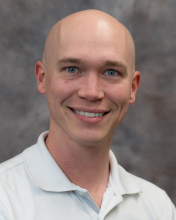 A man with blue eyes in a white shirt against a gray back drop. 