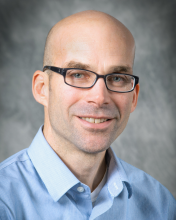 A man with blue yes and glasses against a gray background. 