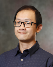 A man with glasses sits against a gray backdrop