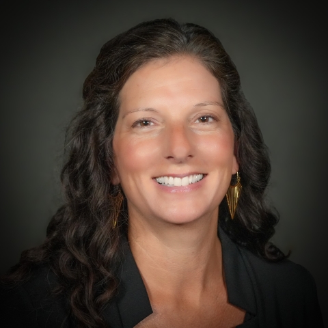A headshot of a woman with brown hair on a gray background. 