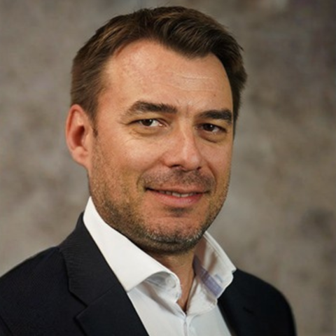 A man with dark hair in a black suit against a gray background. 