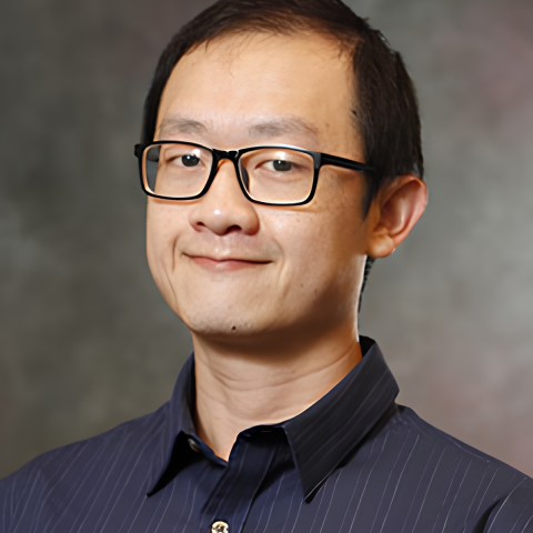 A man with glasses sits against a gray backdrop