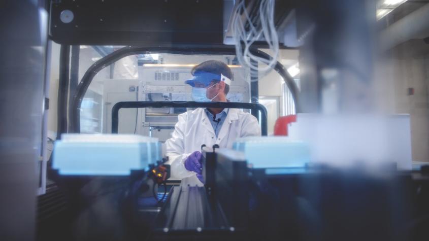 A male worker at the Cornell COVID Testing Lab, seen through stacks of samples; he is wearing a mask, a visor, and gloves