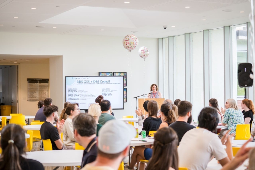 Paula Cohen speaking at a podium in front of groups of people sitting at tables