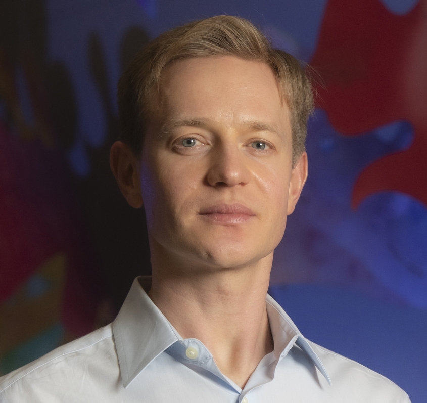 Portrait photograph of Dr. Maksim Plikus in front of a blue and red background.