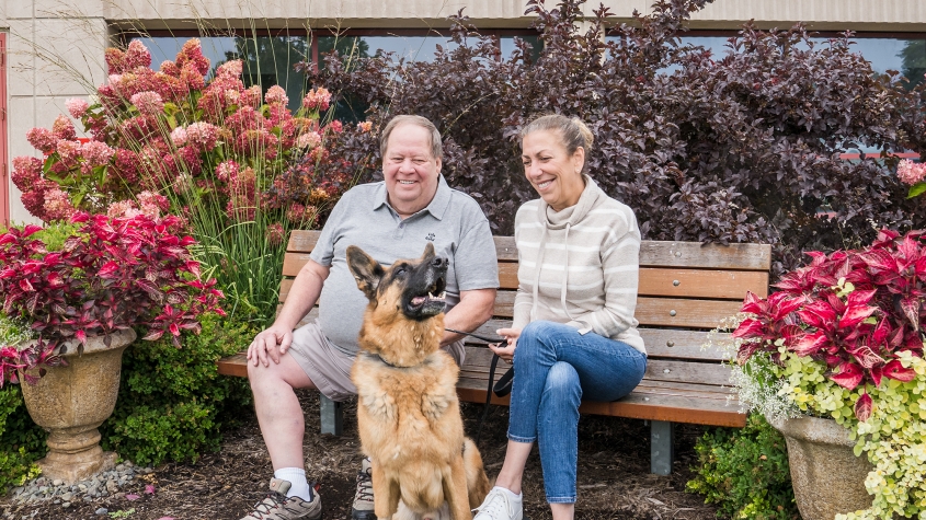 A German shepherd seated in front of a bench, where his owners smile down on him