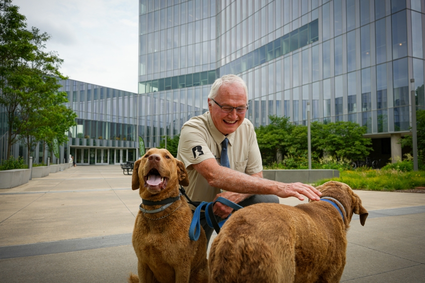 A man with glasses ang gray hair sits with two dogs in front of a large building. 