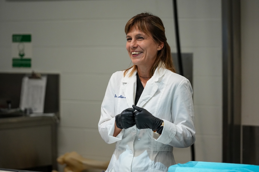 A woman smiling while wearing a white lab coat and black gloves. 
