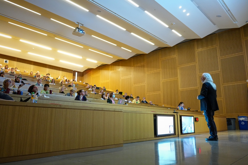 A man with long white hair and wearing a black suit stands in front of a large lecture hall. 