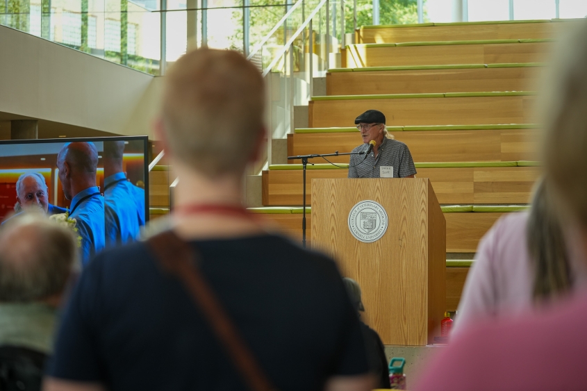 A man with a mustache wearing a black hat speaks at a podium in the CVM atrium.. 