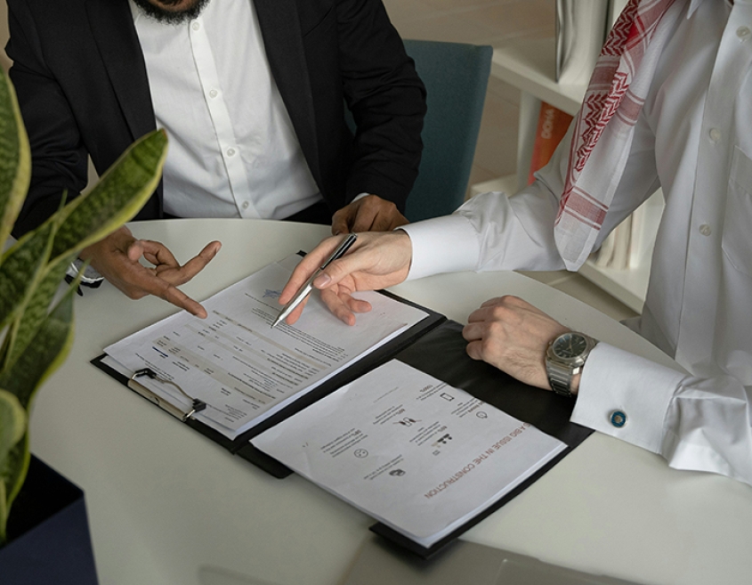 Two individuals at a conference table discuss materials on a clipboard