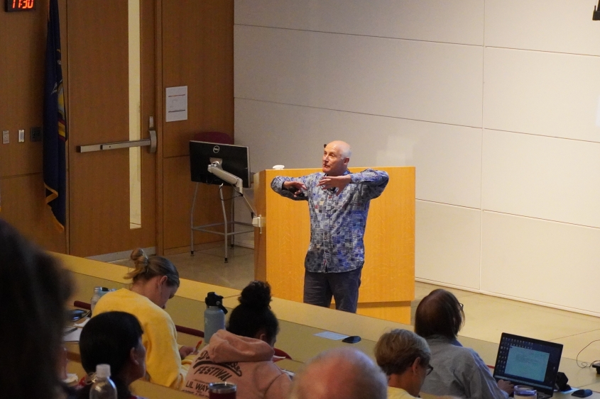 A bald man with a bright, multi-patterned shirt leads a discussion at the front of a lecture hall. 
