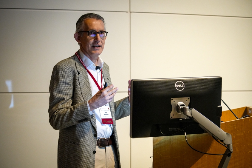 A man with glasses talks in front of a white wall and podium. 