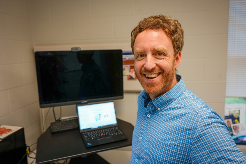 Charles Danko smiling at camera standing in front of computer monitor