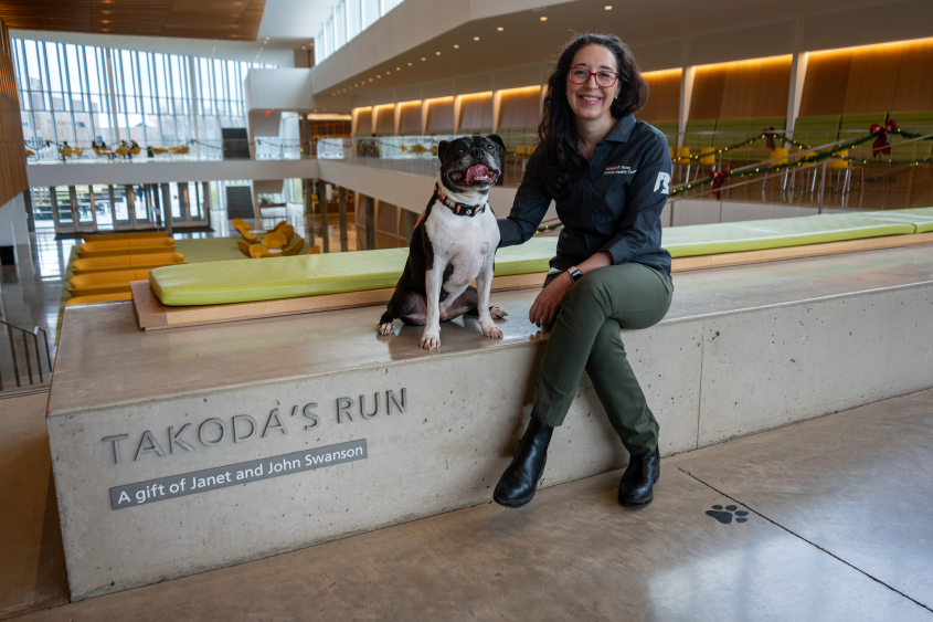 A woman with dark hair and glasses sits in the CVM atrium with a small black dog.